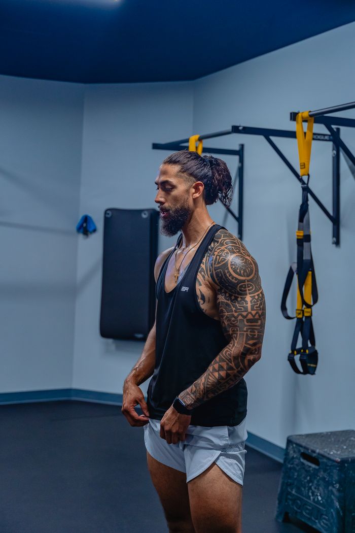Muscular tattooed man in black tank top, posing in an indoor fitness studio with training equipment.